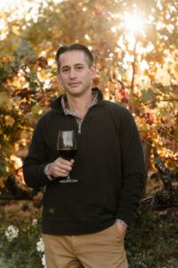 Man standing in front of vines in the fall holding a glass of wine
